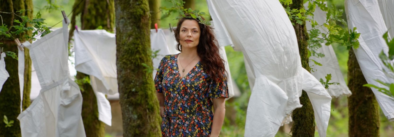 Marthe Vassallo pose en robe longue à fleurs au milieu des arbres. Des vêtements blancs sont étendus sur des cordes à linge autour d'elle.